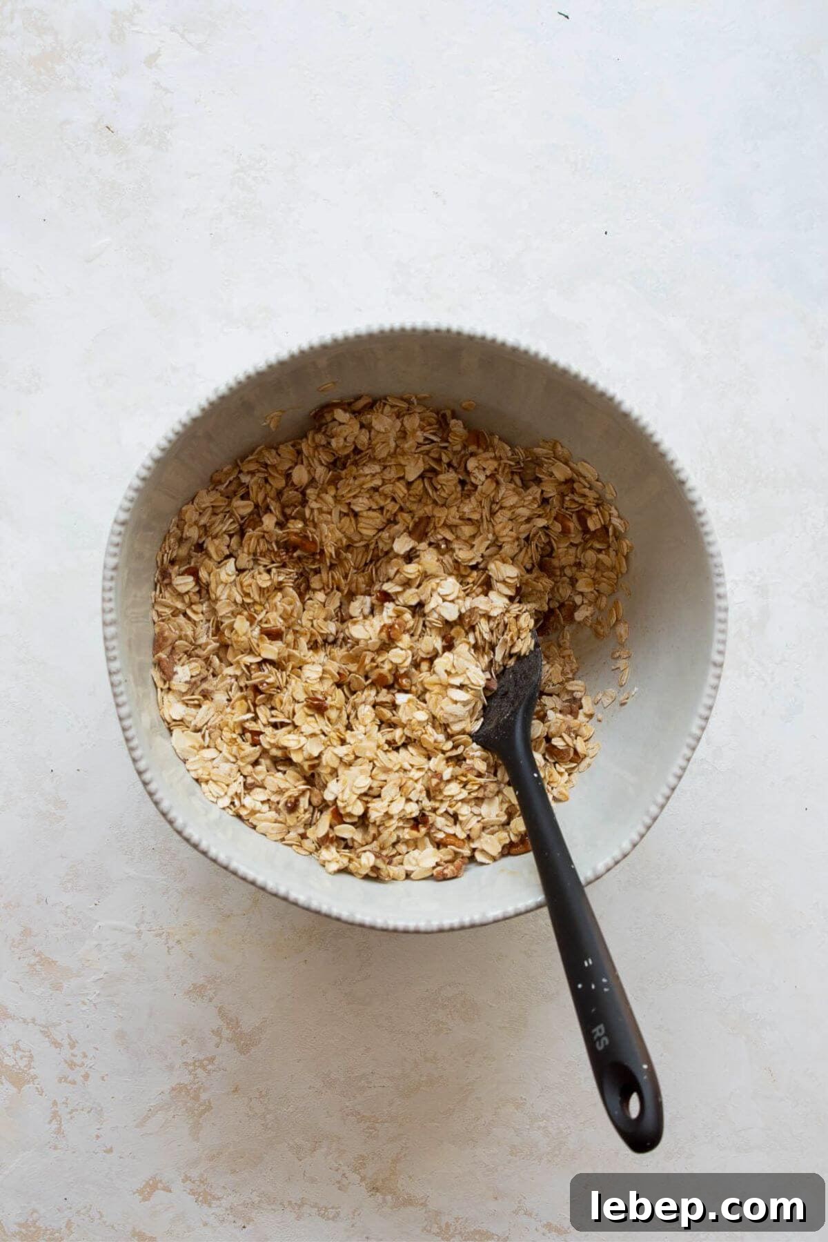 Healthy granola mixture tossed with melted coconut oil, maple syrup, and vanilla in a white bowl, ready to spread on a baking sheet.
