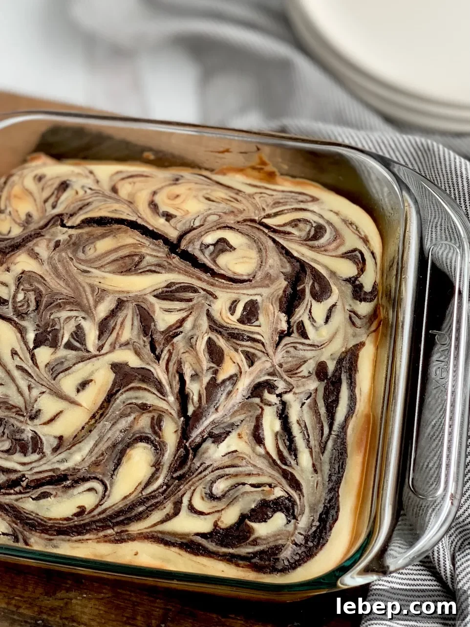Decadent Cheesecake Swirl Brownies — Rich Chocolate & Cream Cheese 6 A glass 8x8-inch baking dish filled with a baked marbled brownie mixture with swirls of cream cheese and dessert plates in the background.