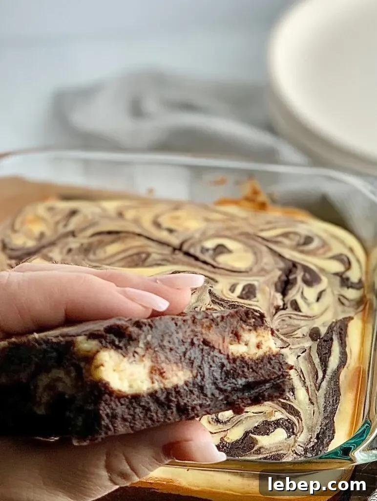 Decadent Cheesecake Swirl Brownies — Rich Chocolate & Cream Cheese 5 A person folding a cut out brownie with chunks of white cream cheese in it with an 8x8-inch baking dish filled with a baked marbled brownie mixture with swirls of cream cheese and dessert plates in the background.