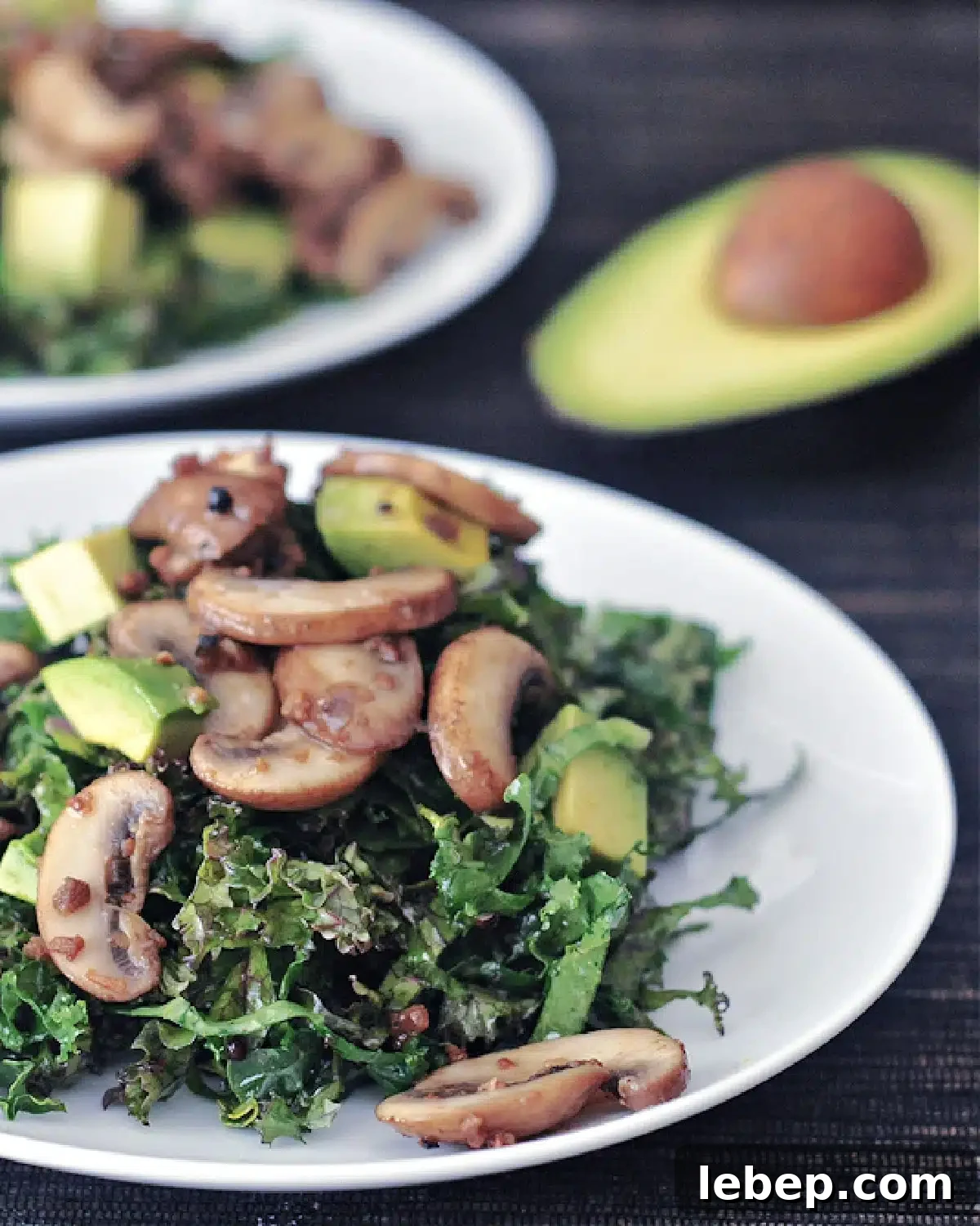 Kale Salad with Warm Bacon Dressing @spabettie #glutenfree #vegan two white plates of kale salad with warm bacon dressing, one plate blurred in background with a half avocado. salad includes kale, cubed avocado, bacon flavored mushrooms
