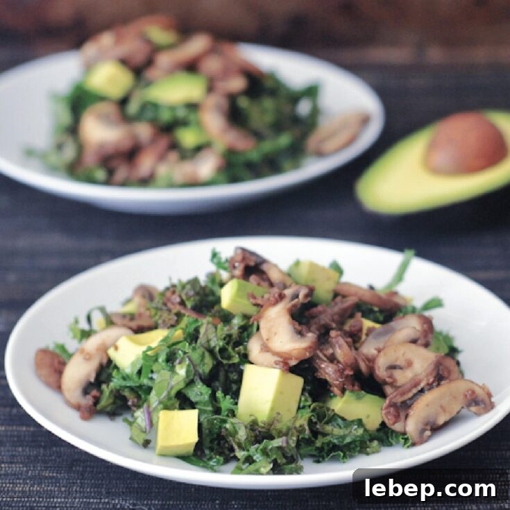 two white plates of kale salad with warm bacon dressing, one plate blurred in background with a half avocado. salad includes kale, cubed avocado, bacon flavored mushrooms