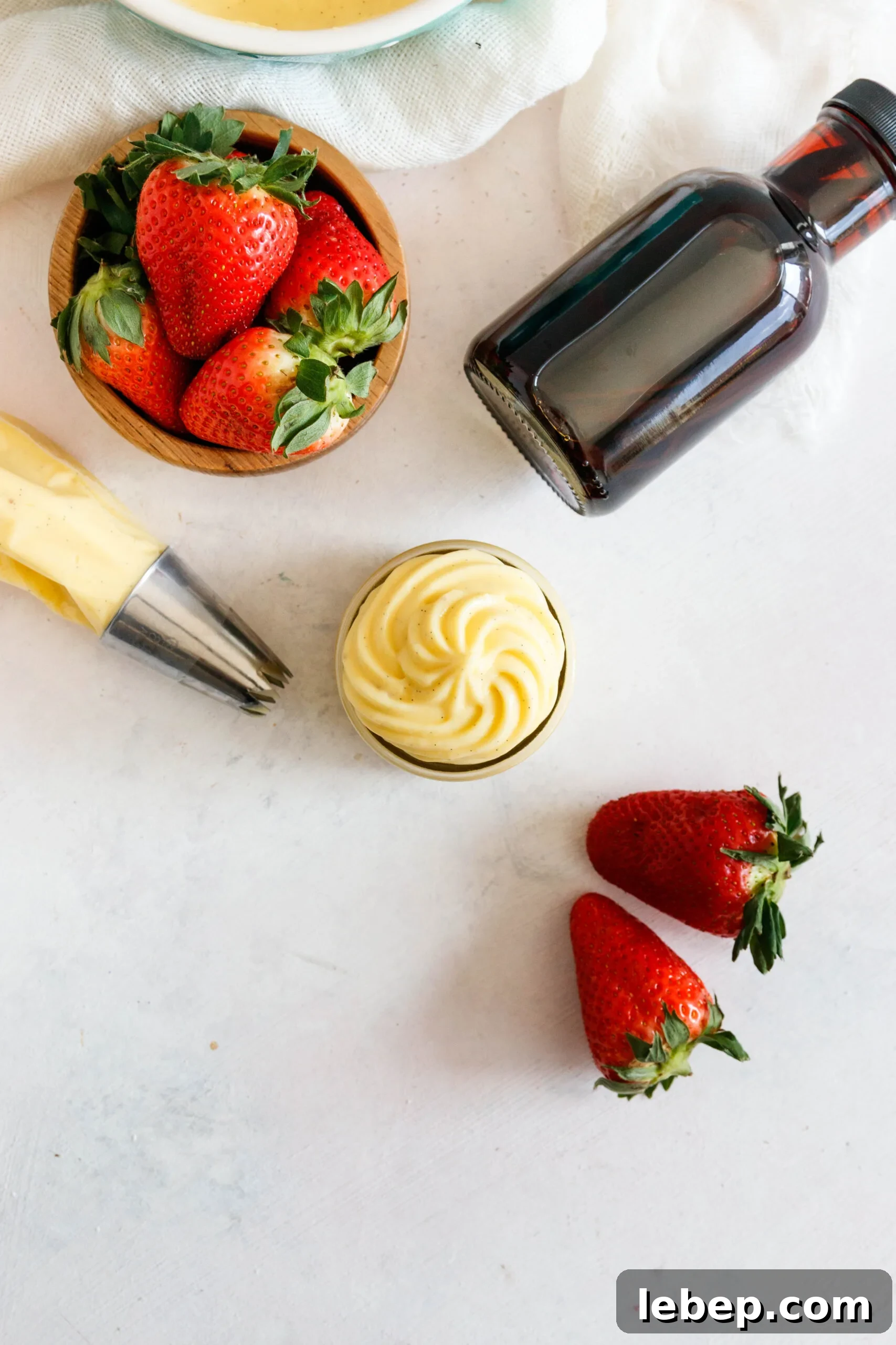 Classic Pastry Cream (Crème Pâtissière) Recipe for Desserts 4 flat lay photo of the pastry cream on a textured white background. Some of the cream is in a pastry bag with a piping tip, the rest is piped into a jar. There's a bottle of extract and some scattered strawberries
