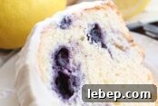 A slice of lemon blueberry bundt cake frosted with lemon cream cheese frosting sitting on a small white plate. Above the plate is a whole lemon and a blueberry lemon bundt cake frosted with lemon cream cheese frosting with a slice taken out of it.