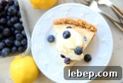In the background are a couple of lemons, two forks, and a container of blueberries. There's a white dessert plate with a slice of lemon blueberry pie topped with whipped cream, candied lemon zest and a few blueberries. On each side of the slice of pie is a single blueberry.