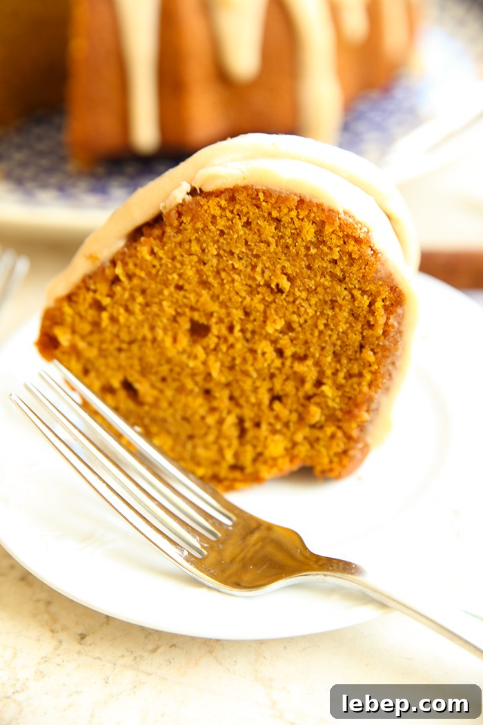 A slice of pumpkin spice cake on a small white plate with a fork. Faded in the background is a pumpkin bundt cake with a slice cut out of it.