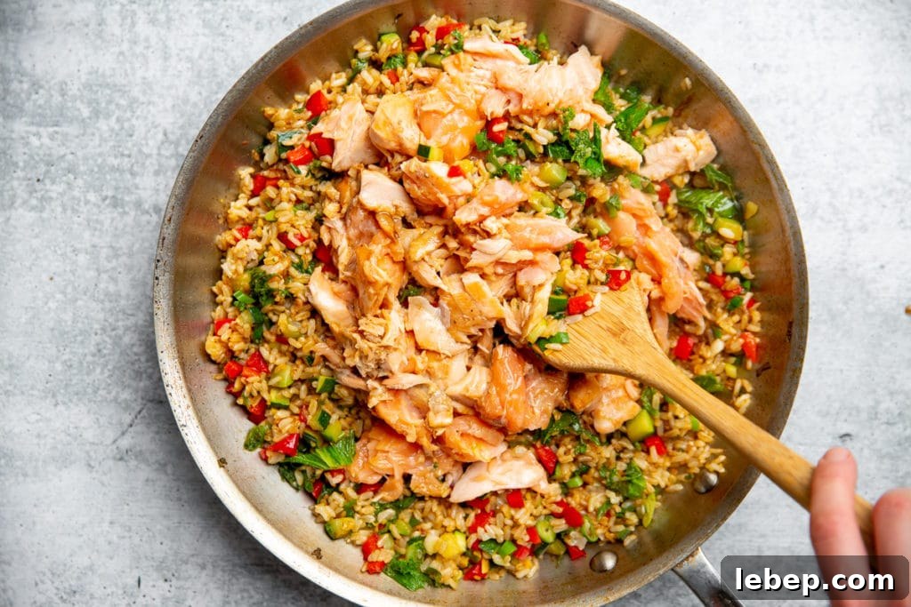 Process shot showing a hand stirring the cooked salmon into the fried rice with a wooden spoon. 