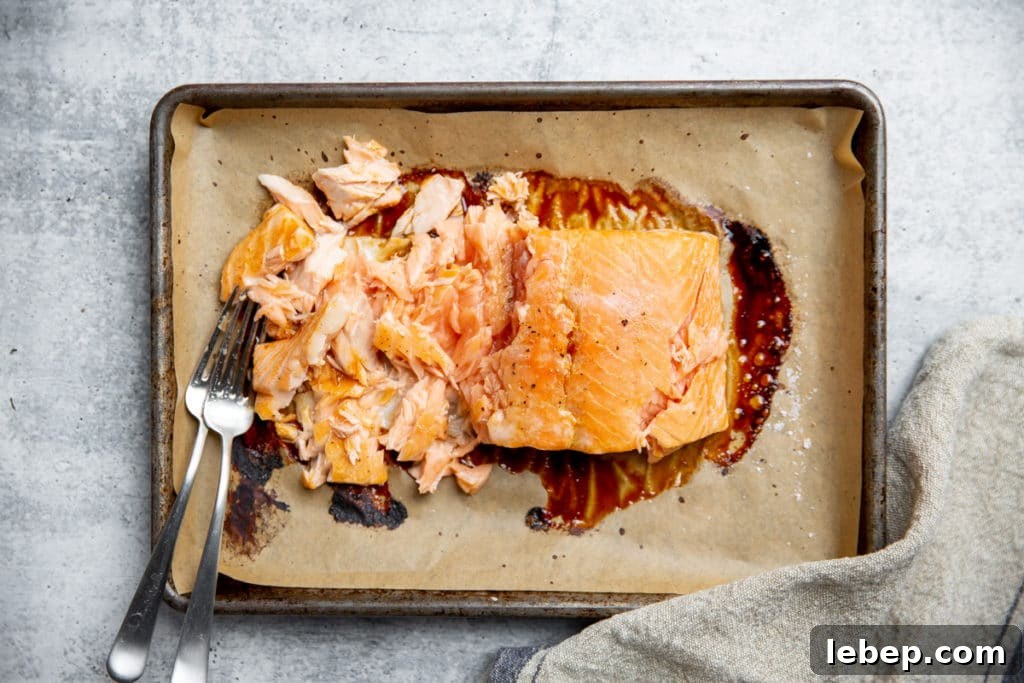 Process shot showing baked salmon on a parchment lined baking sheet, being flaked with two forks. 