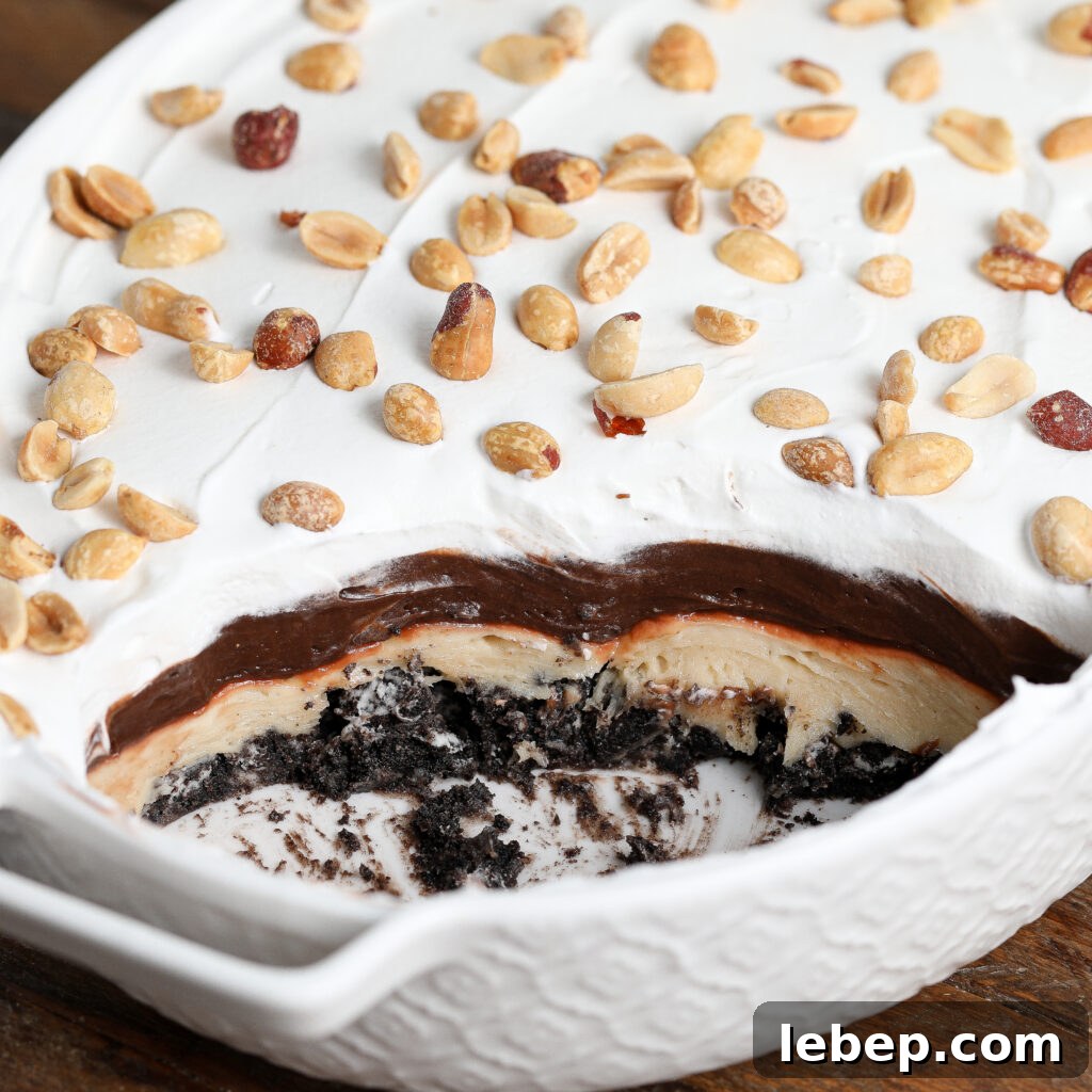 A dessert in a white dish with layers of chocolate, cream, and cookie crumbs, topped with whipped topping and salted peanuts; a portion has been served to reveal the inside of this decadent Gluten and Dairy Free Oreo Peanut Butter Pie.