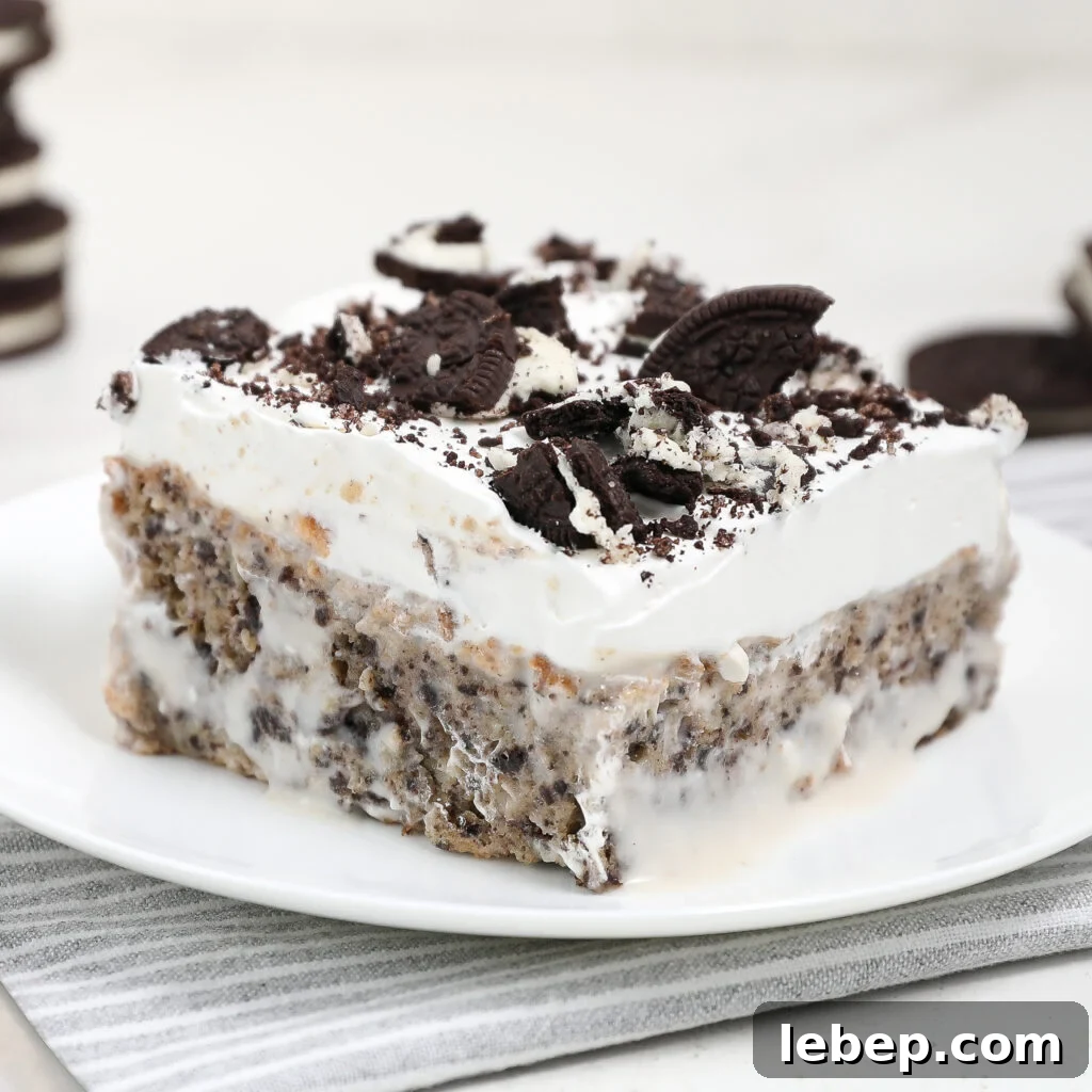 A slice of cookies and cream icebox cake, inspired by Oreo tres leches, topped with whipped cream and crushed chocolate sandwich cookies sits on a white plate with a gray-striped napkin underneath.