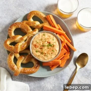 Obatzda german cheese spread in a bowl topped with paprika and chives, surrounded by soft pretzels and carrot sticks, two glasses of beer are in the upper right.