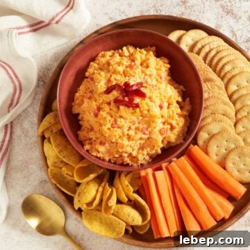 Pimento cheese dip in a red bowl on a round wooden tray, surrounded by butter crackers, carrot sticks, and corn chips. A gold spoon is on the lower left, a red and white striped towel is on the upper left.