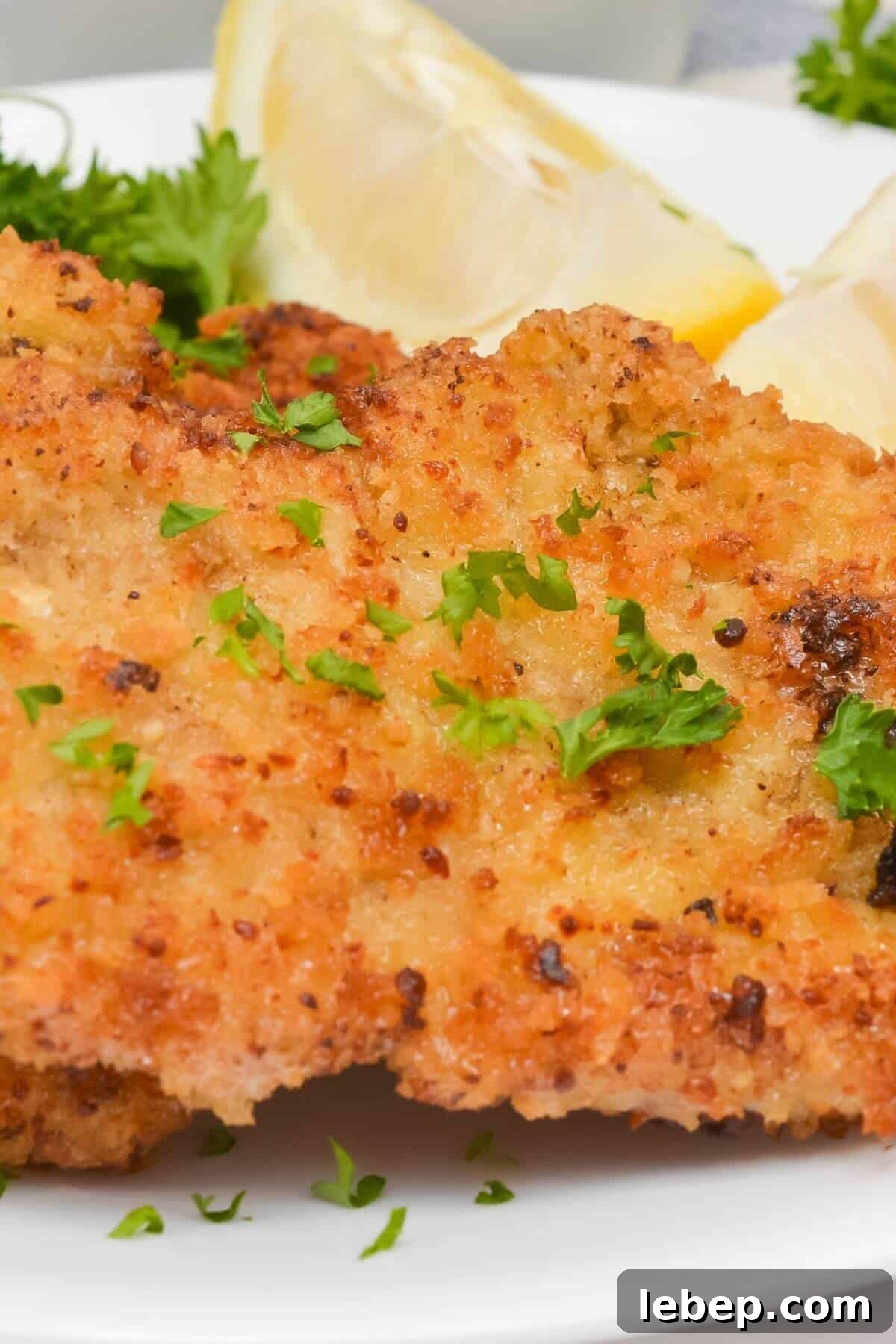 Close-up of breaded and fried pork cutlets, reminiscent of a classic pork cutlet, garnished with chopped parsley and served with lemon wedges on a white plate.