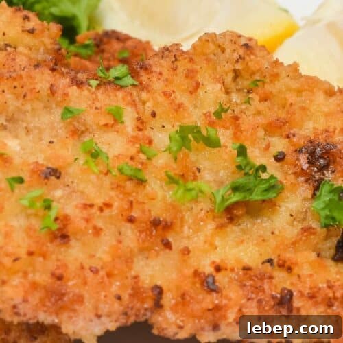 Close-up of breaded and fried pork cutlets, reminiscent of a classic pork cutlet, garnished with chopped parsley and served with lemon wedges on a white plate.
