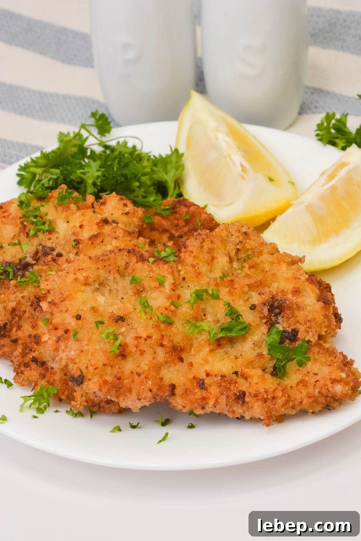 Two thinly pounded and breaded meat  garnished with chopped parsley and served with lemon wedges and parsley sprigs on a white plate.