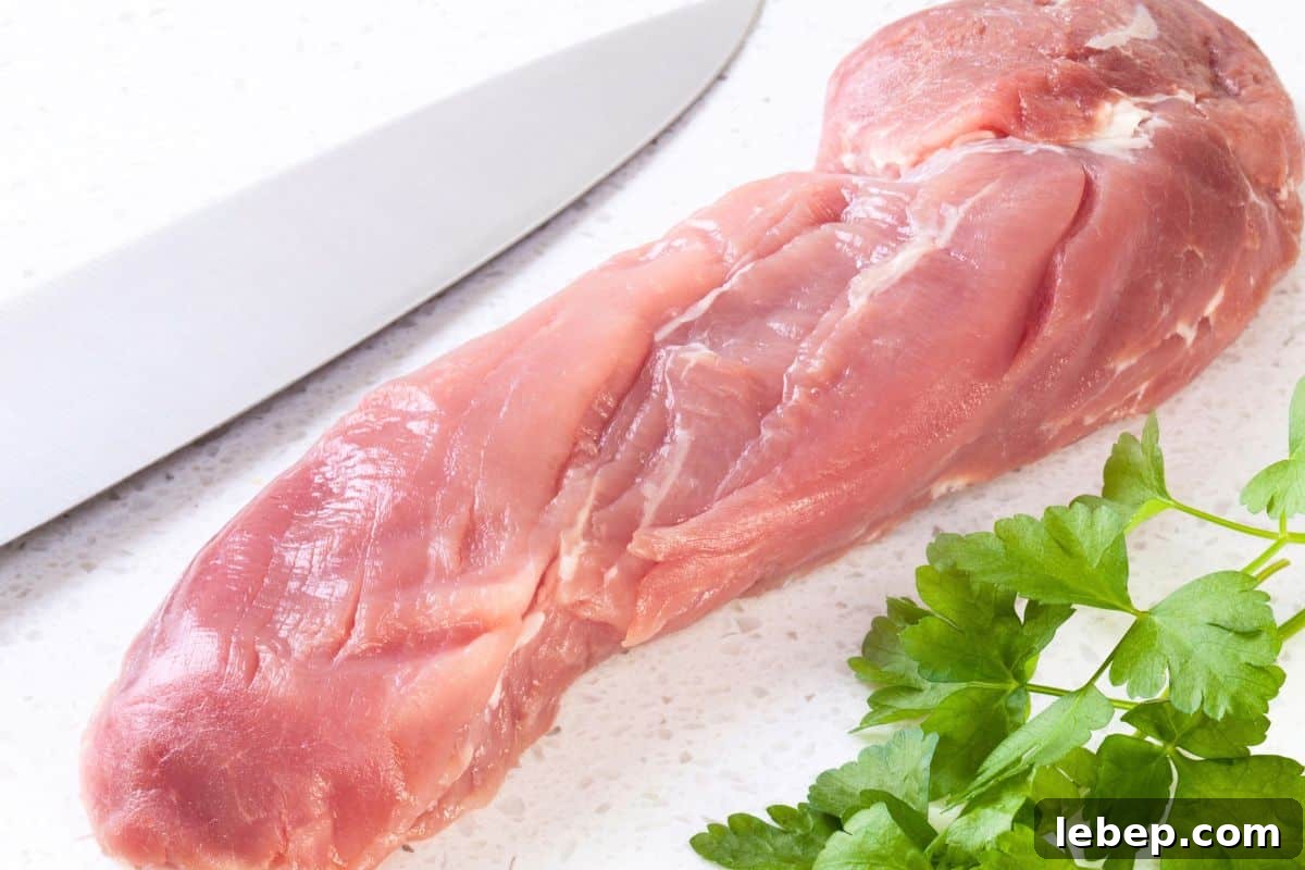 Raw pork tenderloin on a white surface next to a kitchen knife and a sprig of fresh parsley, ideal for slicing into tender pork cutlets.