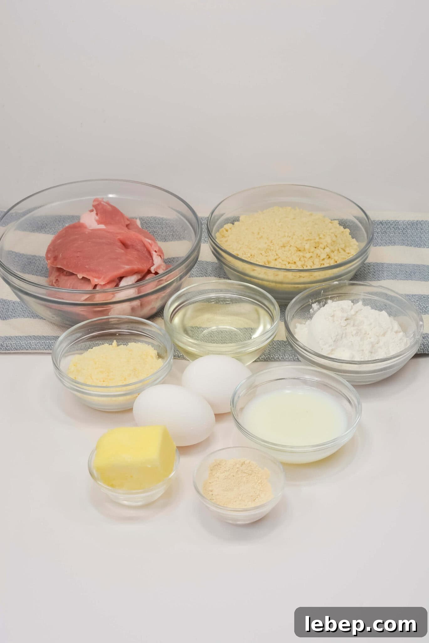 Assorted ingredients in glass bowls, including raw pork cutlet, eggs, flour, grated cheese, breadcrumbs, oil, butter, milk, and a light-colored powder, arranged on a striped towel.
