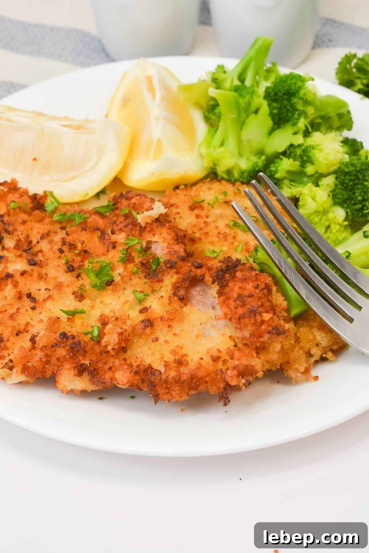 A plate with breaded cutlet, steamed broccoli, lemon wedges, and a fork resting on the side.