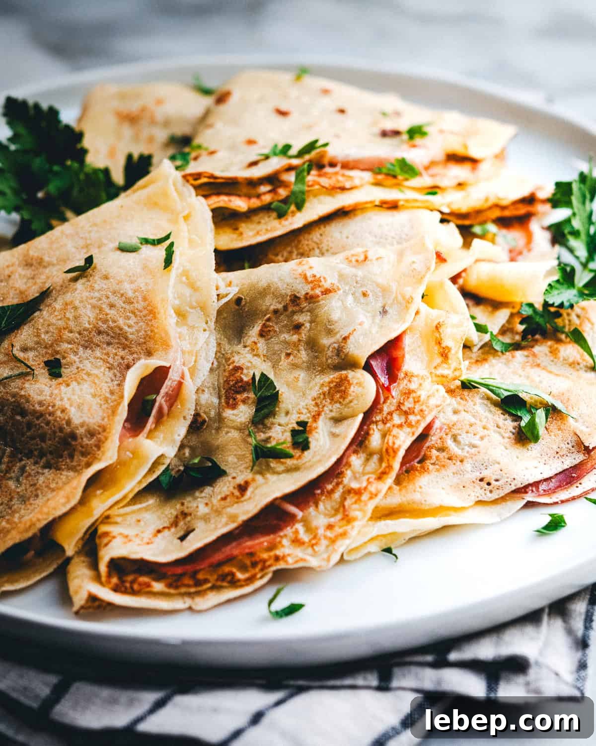 Savory Crepes: 10 Flavorful Fillings and Cooking Tips 2 A plate filled with freshly made crepes folded in quarters, garnished with chopped herbs. The crepes are lightly browned and appear stuffed with ingredients such as meat and cheese. The plate rests on a striped cloth napkin.
