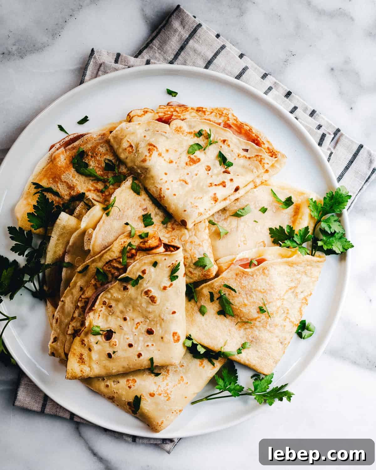 Savory Crepes: 10 Flavorful Fillings and Cooking Tips 4 A white plate filled with neatly folded crepes garnished with chopped fresh parsley. The plate is set on a gray and white striped napkin, all placed on a marble countertop.