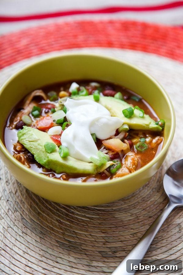 a bowl of taco soup topped with avocado and sour cream in a green bowl on a wicker placemat. 