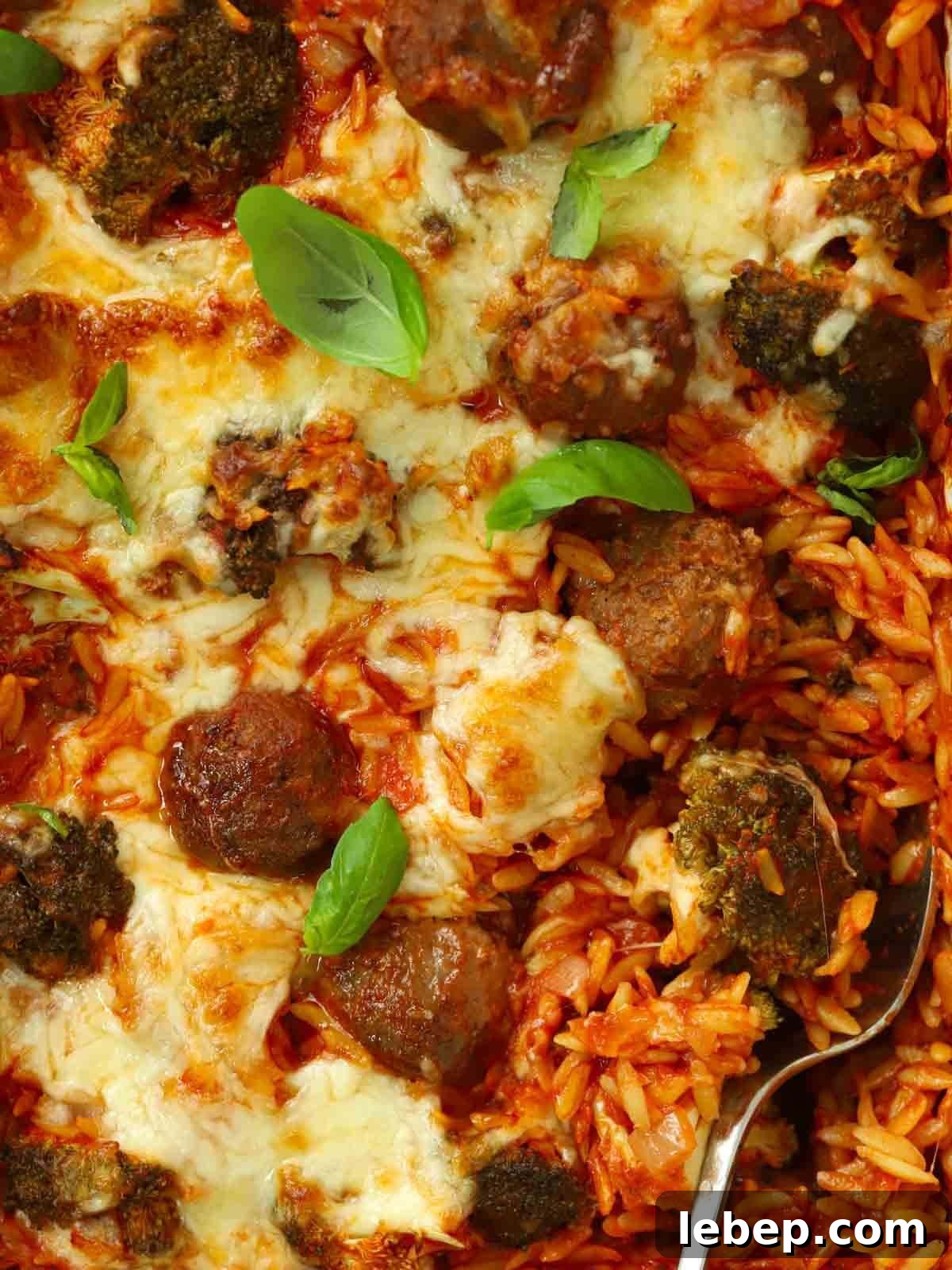 A close up of meatballs, orzo and cheese, cooked in one dish together in the oven. A spoon is showing, ready to serve.