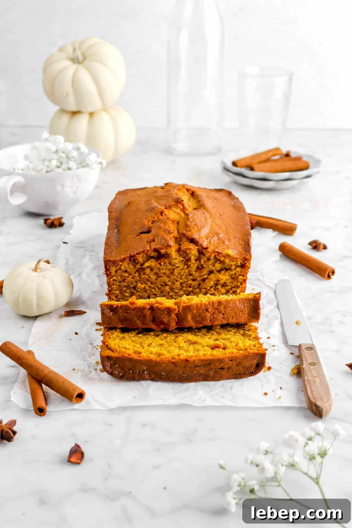 Moist Pumpkin Bread Recipe with Warm Spices 2 pumpkin bread on parchment paper with whole spices, two slices laying in front, a knife, flowers, and pumpkins