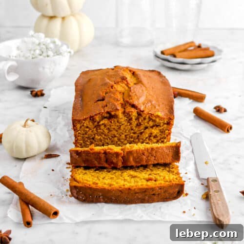 Moist Pumpkin Bread Recipe with Warm Spices 20 pumpkin bread on parchment paper with whole spices, two slices laying in front, a knife, flowers, and pumpkins