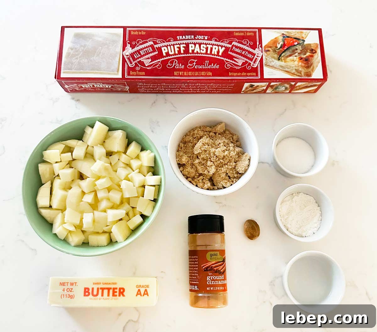 A kitchen counter with puff pastry, chopped apples, brown sugar, butter, cinnamon, sugar, flour, water and nutmeg.