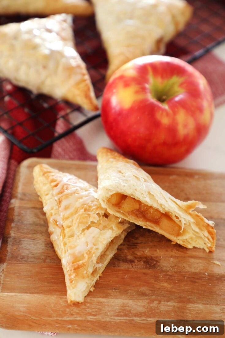 Apple turnovers on a cutting board with an apple and turnovers cooling on a baking rack.