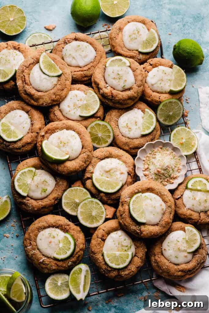 Key Lime Pie Cookies with Make-Ahead Prep Tips 8 Key lime pie cookies on a cooling rack, on a blue backdrop. The cookies are brightly and beautifully decorated with lime slices.