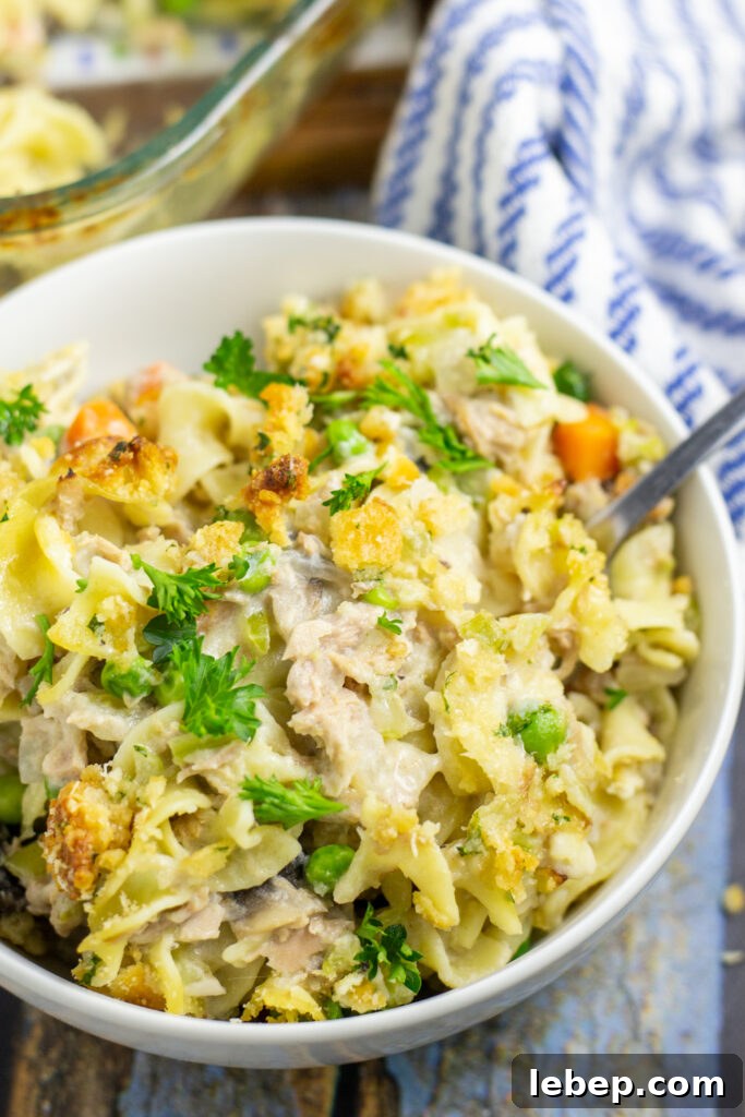 Creamy Tuna Noodle Casserole Recipe with Crispy Topping 2 Tuna Noodle Casserole in a white bowl with a fork sticking out on a rustic wood background with a linen napkin and the full glass casserole dish behind.