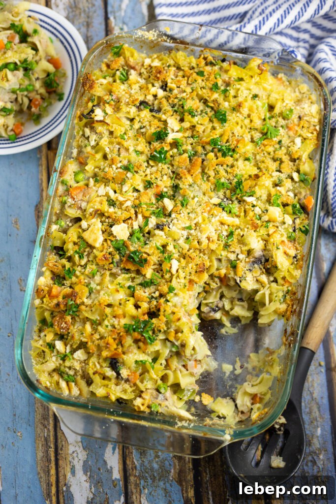 Creamy Tuna Noodle Casserole Recipe with Crispy Topping 3 Overhead view of tuna noodle casserole in a glass casserole dish topped with fresh parsley on a rustic wood background with a linen napkin and a small plate with a scoop of the casserole on it.