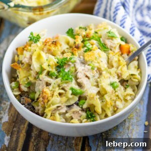 Tuna Noodle Casserole in a white bowl with a fork sticking out on a rustic wood background with a linen napkin and the full glass casserole dish behind.