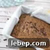 Dairy-free chocolate zucchini bread in a baking loaf with parchment paper and a blue linen and scoop of collagen powder around it.