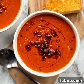 overhead shot of red pepper soup served in white bowl on wooden board with spoon and grilled cheese