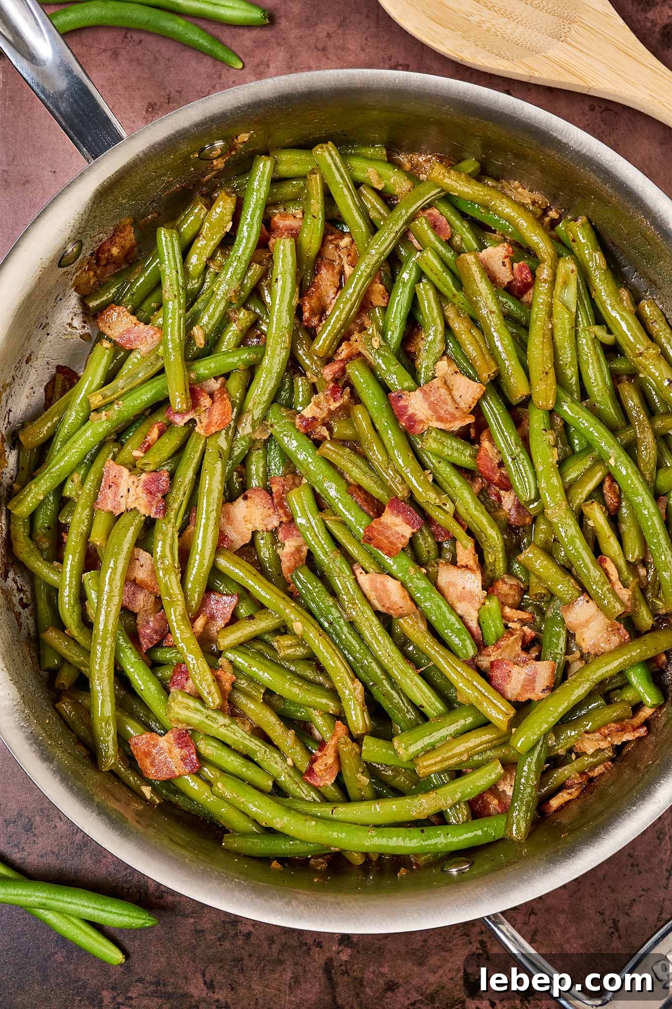 Southern-Style Green Beans with Bacon and Garlic 2 A sauté pan filled with cooked Southern style green beans with bacon, next to a wooden spoon and several scattered fresh beans.