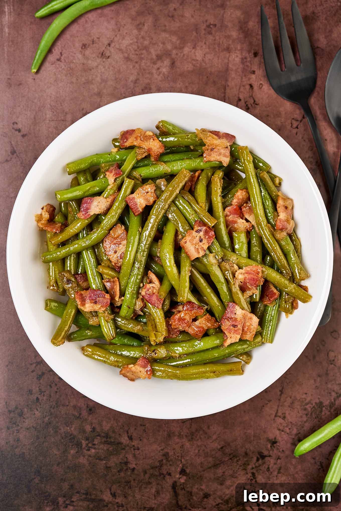 Southern-Style Green Beans with Bacon and Garlic 6 A round, white serving bowl of Southern style green beans with bacon on a rustic brown surface with a black serving fork to the right.