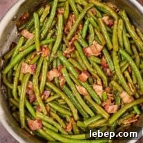 Southern-Style Green Beans with Bacon and Garlic 7 A sauté pan filled with cooked Southern style green beans with bacon, next to a wooden spoon and several scattered fresh beans.
