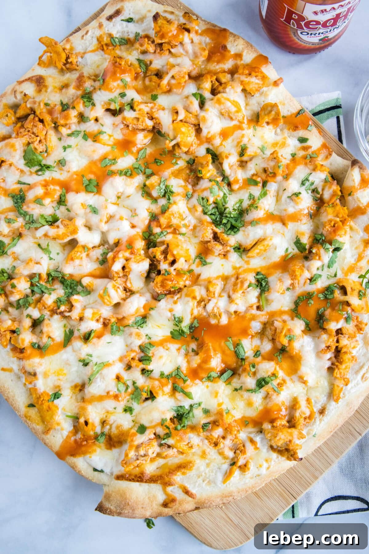 Homemade buffalo chicken pizza is loaded with spicy and juicy chicken, creamy ranch dressing, and melted cheese on a golden-brown crust. Overhead view of a whole buffalo chicken pizza next to a bottle of wing sauce