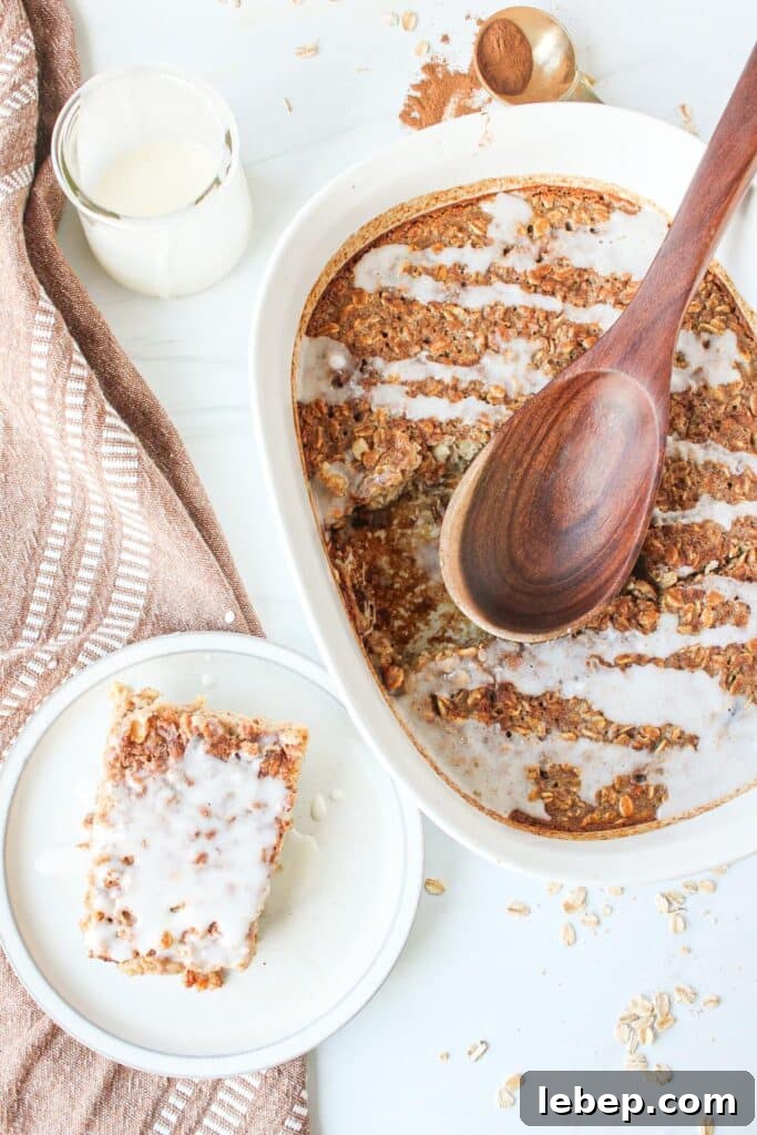 Cinnamon Roll Baked Oatmeal — Morning Bake with Creamy Glaze 2 A white casserole dish with cinnamon roll oatmeal bake drizzled with a cinnamon roll glaze with a slice next to it on a plate.