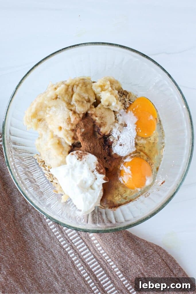 Cinnamon Roll Baked Oatmeal — Morning Bake with Creamy Glaze 4 A clear bowl full of ingredients for an oatmeal bake: banana, Greek yogurt, eggs, cinnamon, and maple syrup unmixed.
