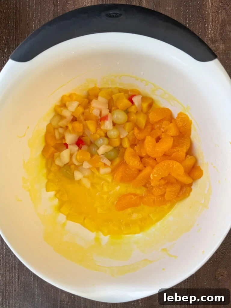 White mixing bowl of pineapple set up in pudding with a can of fruit cocktail and mandarin oranges poured on top.