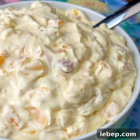 Close up cherry, pineapple tidbit, grape and other fruit cocktail ingredients coated with creamy vanilla pudding mixture heaped up in a white bowl.