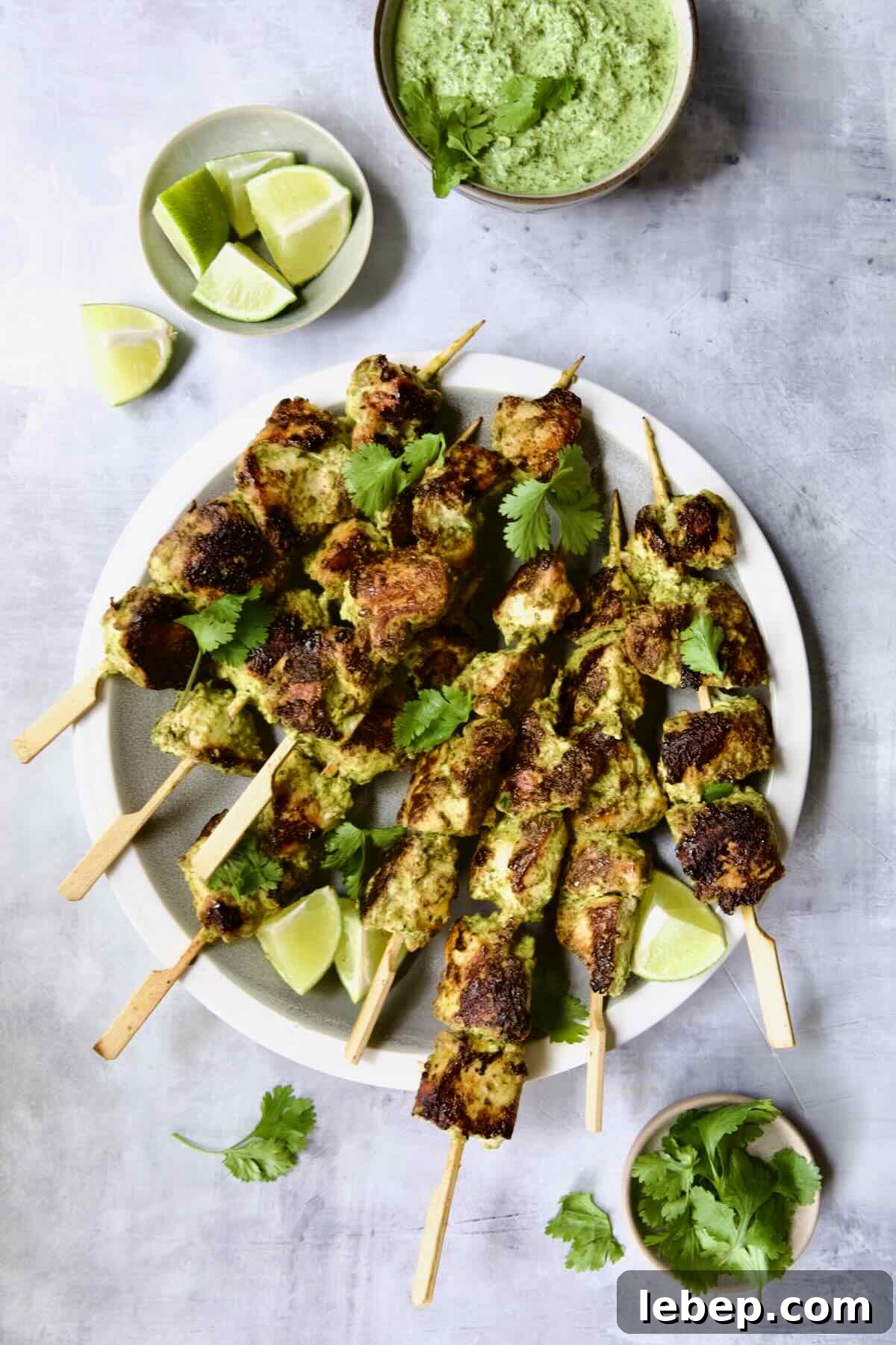 Yogurt Marinated Grilled Chicken Skewers on white plate with Cilantro Mint Chutney, lime wedges and cilantro sprigs in small bowls.