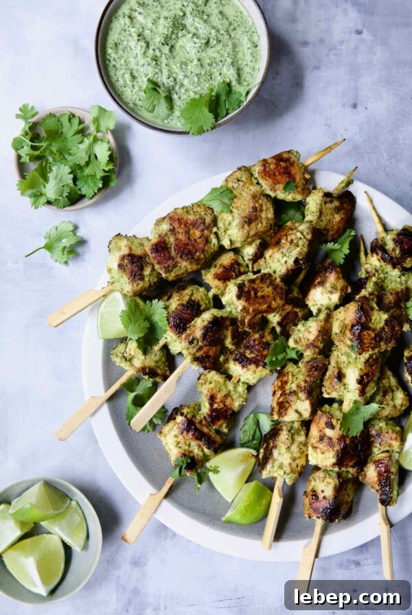 Yogurt Marinated Grilled Chicken Skewers on white plate with Cilantro Mint Chutney, cilantro sprigs and lime wedges in small bowls.