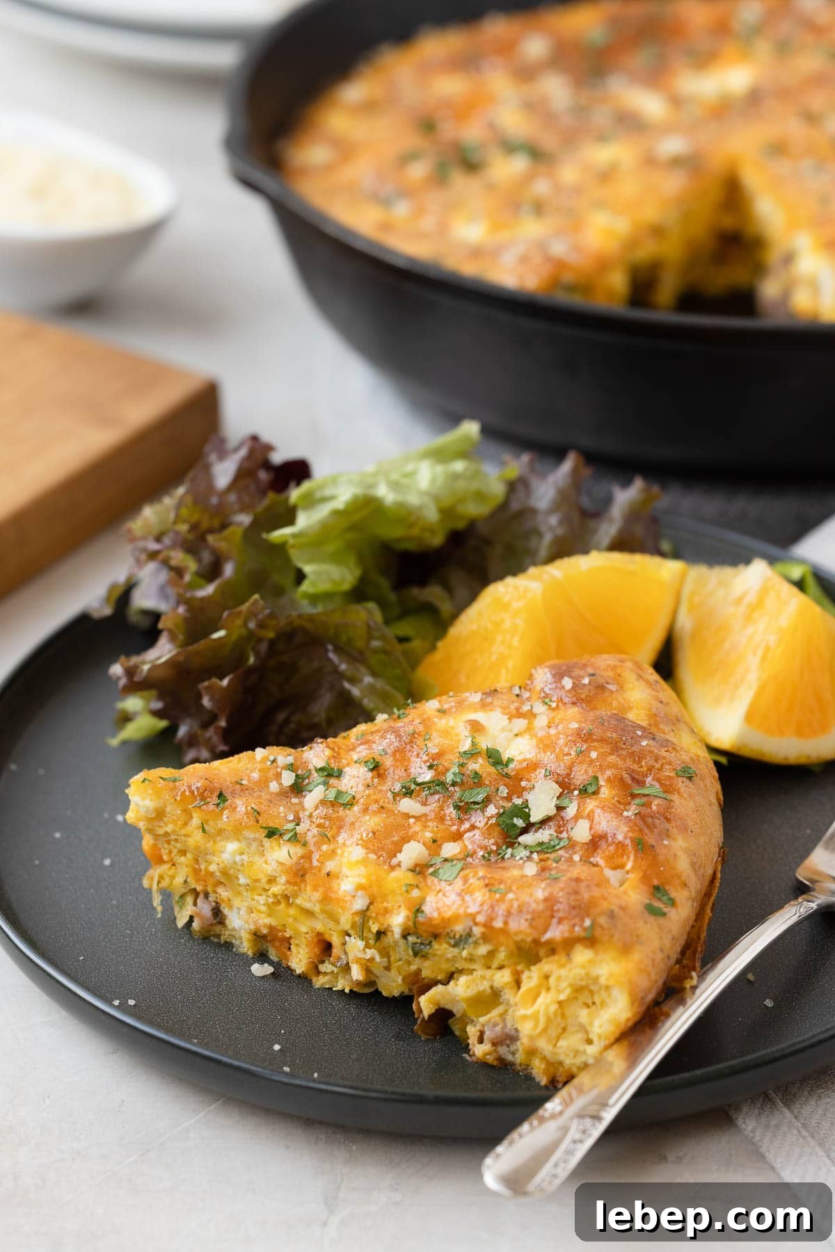 A slice of ham and cheese frittata on a plate with orange wedges and salad.