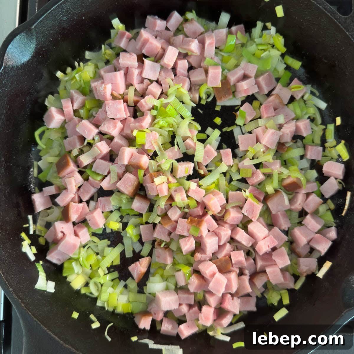 Pink diced ham steak added to a skillet of sauteing leeks for a frittata.