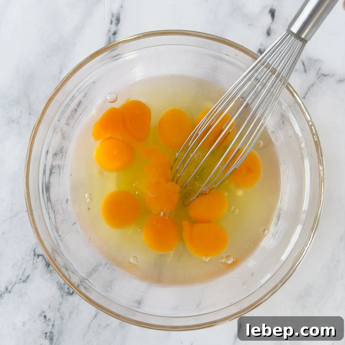 A glass bowl of eggs white and yolks with a whisk on a marble counter.