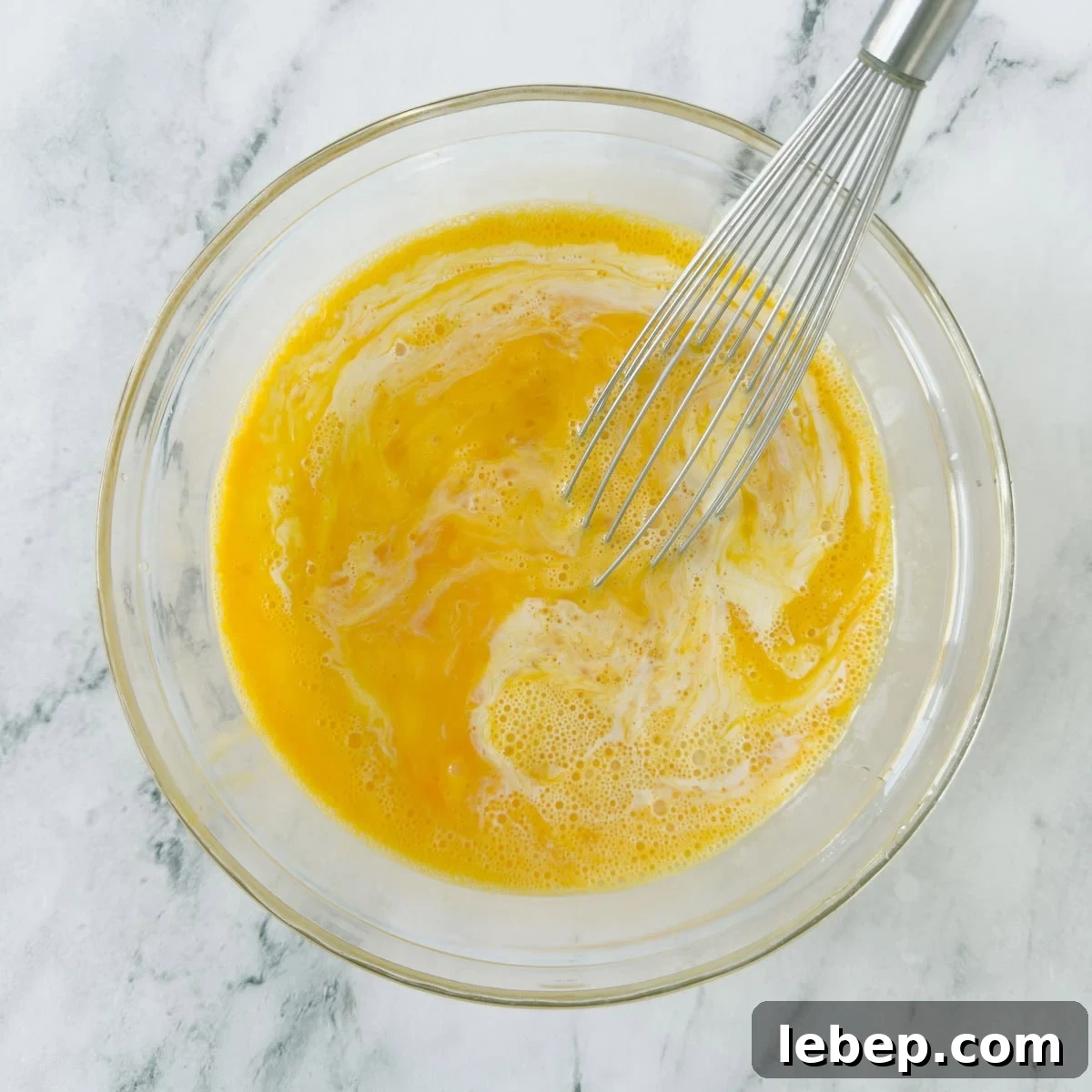 Whisking together eggs and half and half for a custard in a glass bowl with silver whisk.