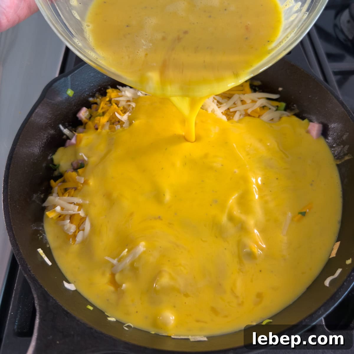 Pouring whisked eggs over vegetables and cheese in a black fry pan.